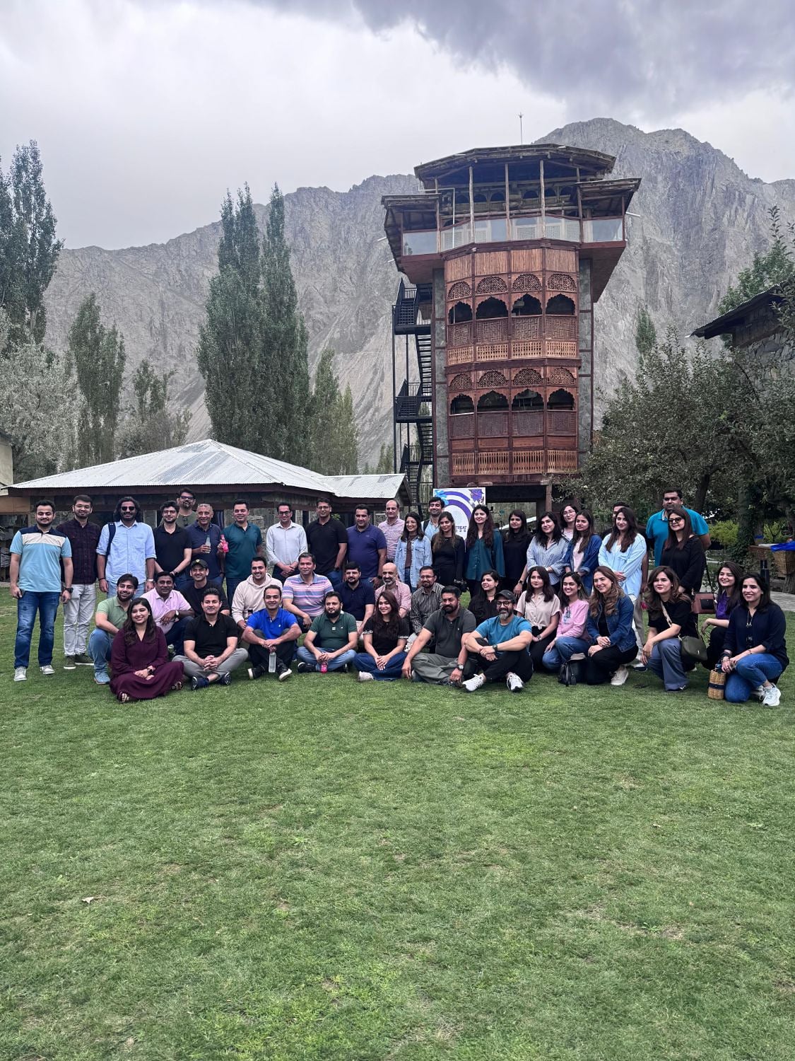 About fifty people from a company retreat posing on the lawn of PC Skardu, in front of its carved wooden tower and rocky mountains