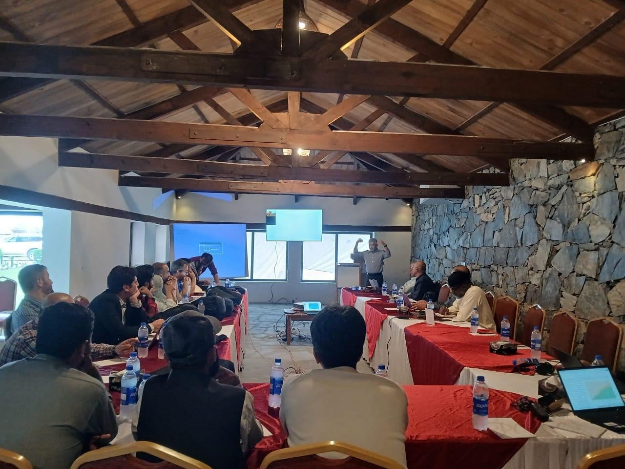 A workshop in a meeting room with a wooden-beamed ceiling and a stone wall, participants at long tables listening to a speaker