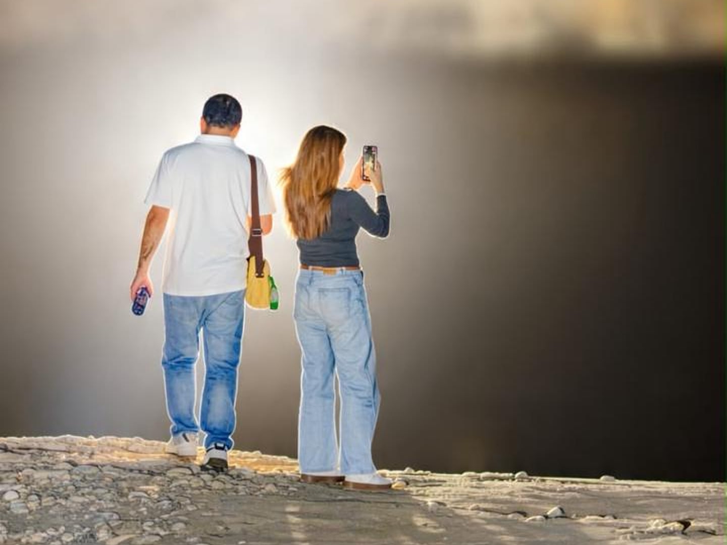 A couple seen from behind at night in Sarfaranga, near Skardu, standing on stony ground in a glow of light; the woman is taking a photo with her phone