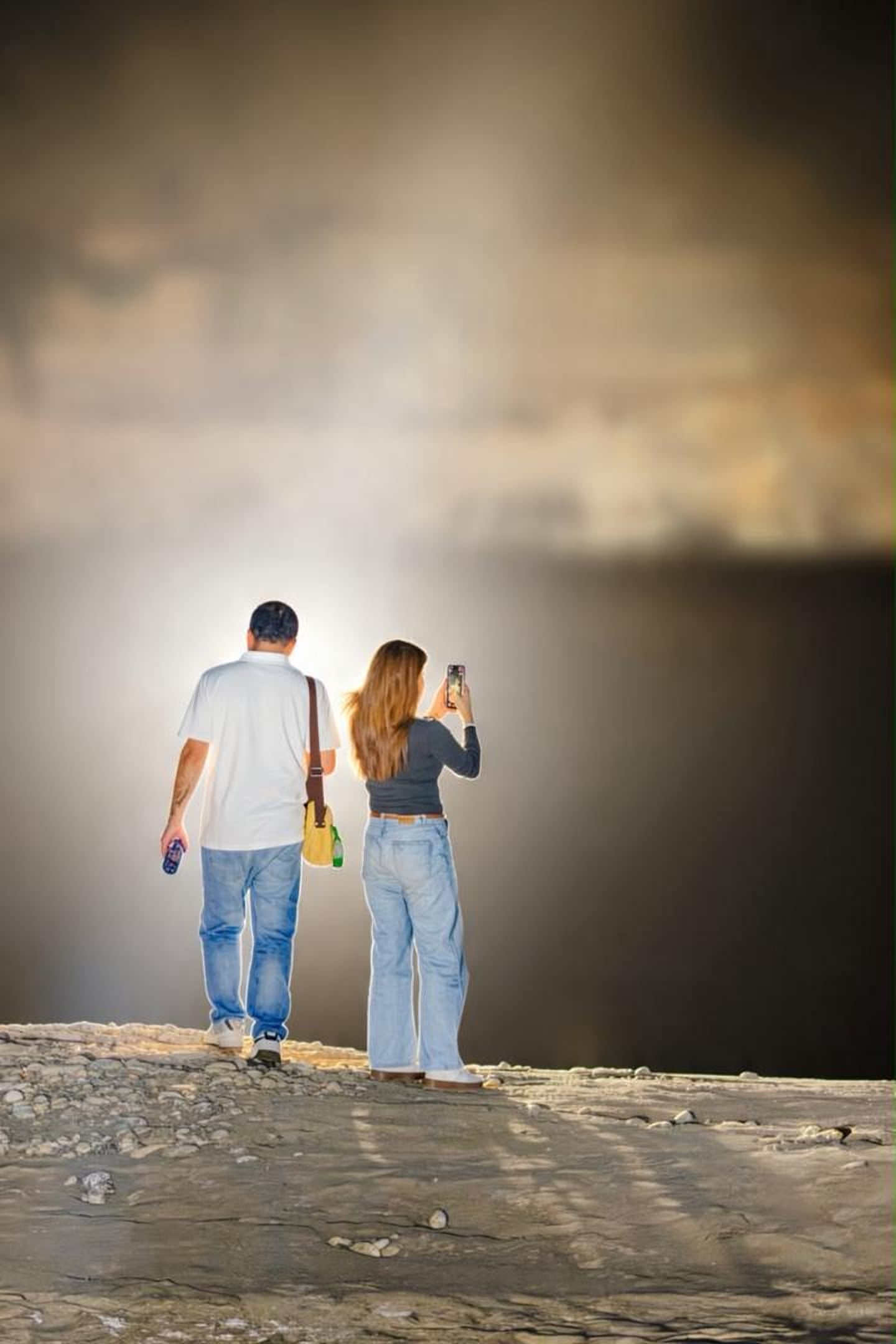 A couple seen from behind at night in Sarfaranga, near Skardu, standing on stony ground in a glow of light; the woman is taking a photo with her phone