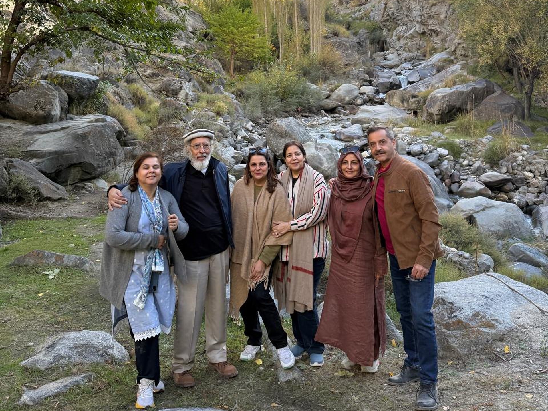 A family of six standing together beside a rocky mountain stream, with golden poplars behind them