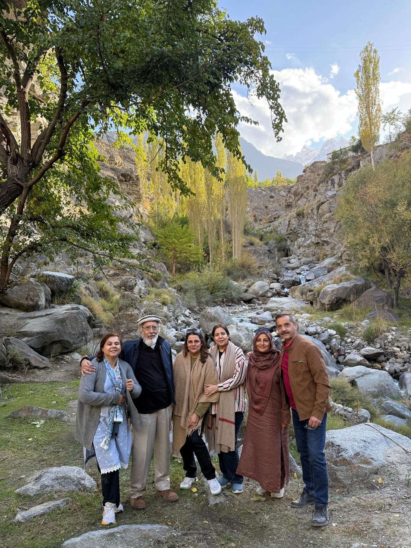 A family of six standing together beside a rocky mountain stream, with golden poplars and snow peaks behind them