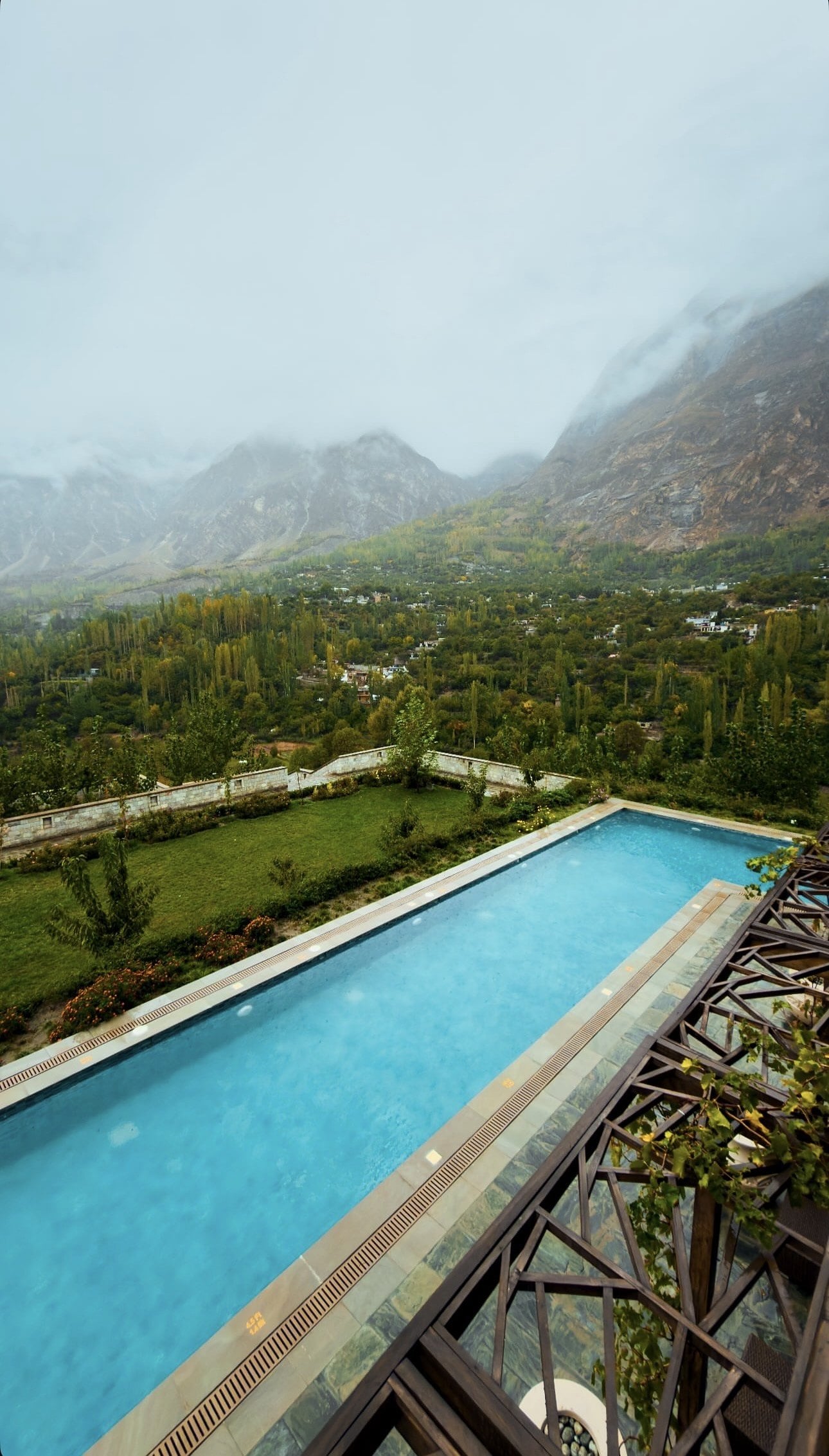 A long blue swimming pool on a terrace above the Hunza valley, with autumn poplars, villages and mist-covered mountains