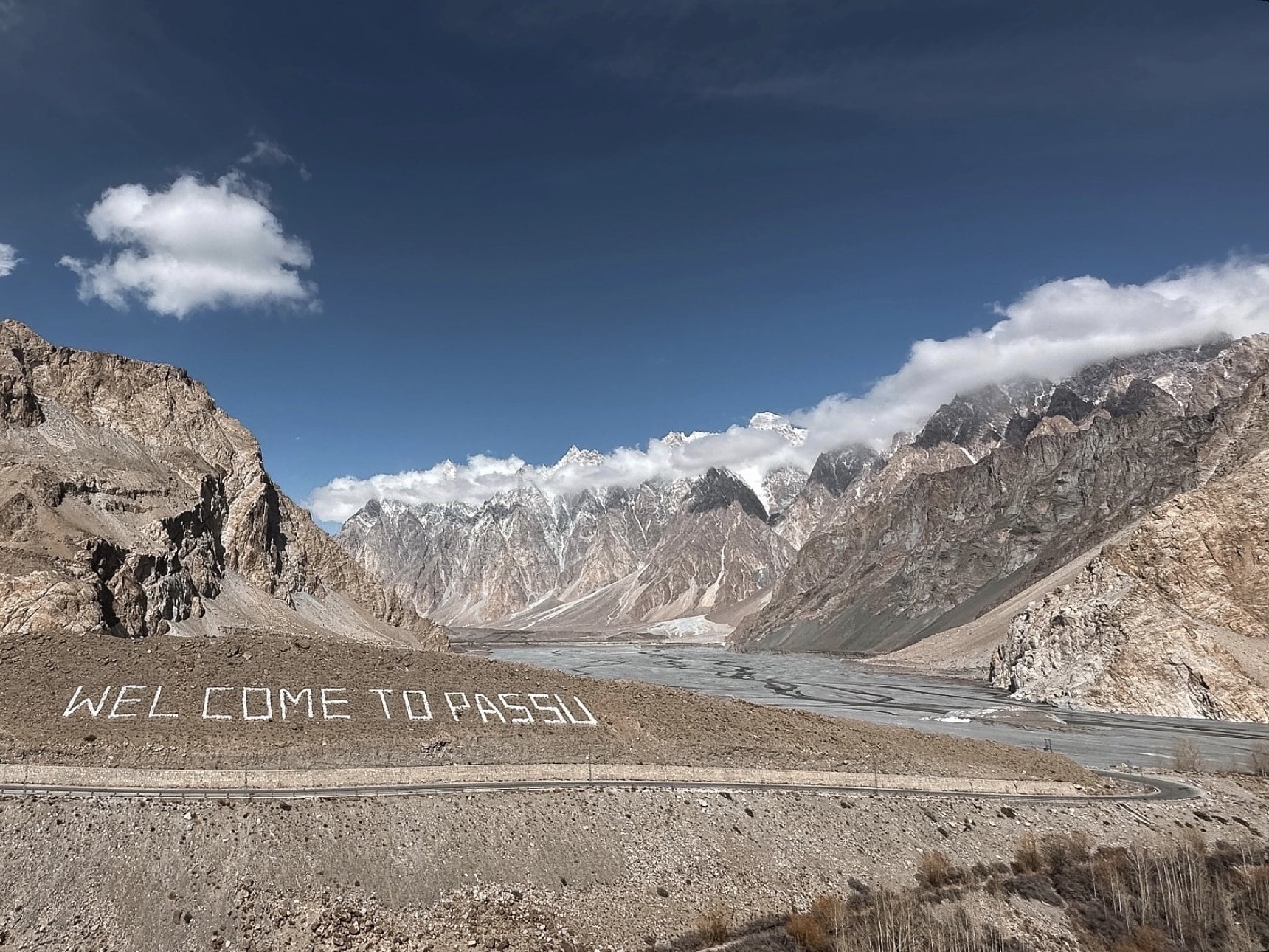 "Welcome to Passu" written in white stones on a hillside, with the jagged Passu Cones and a wide river bed under a blue sky