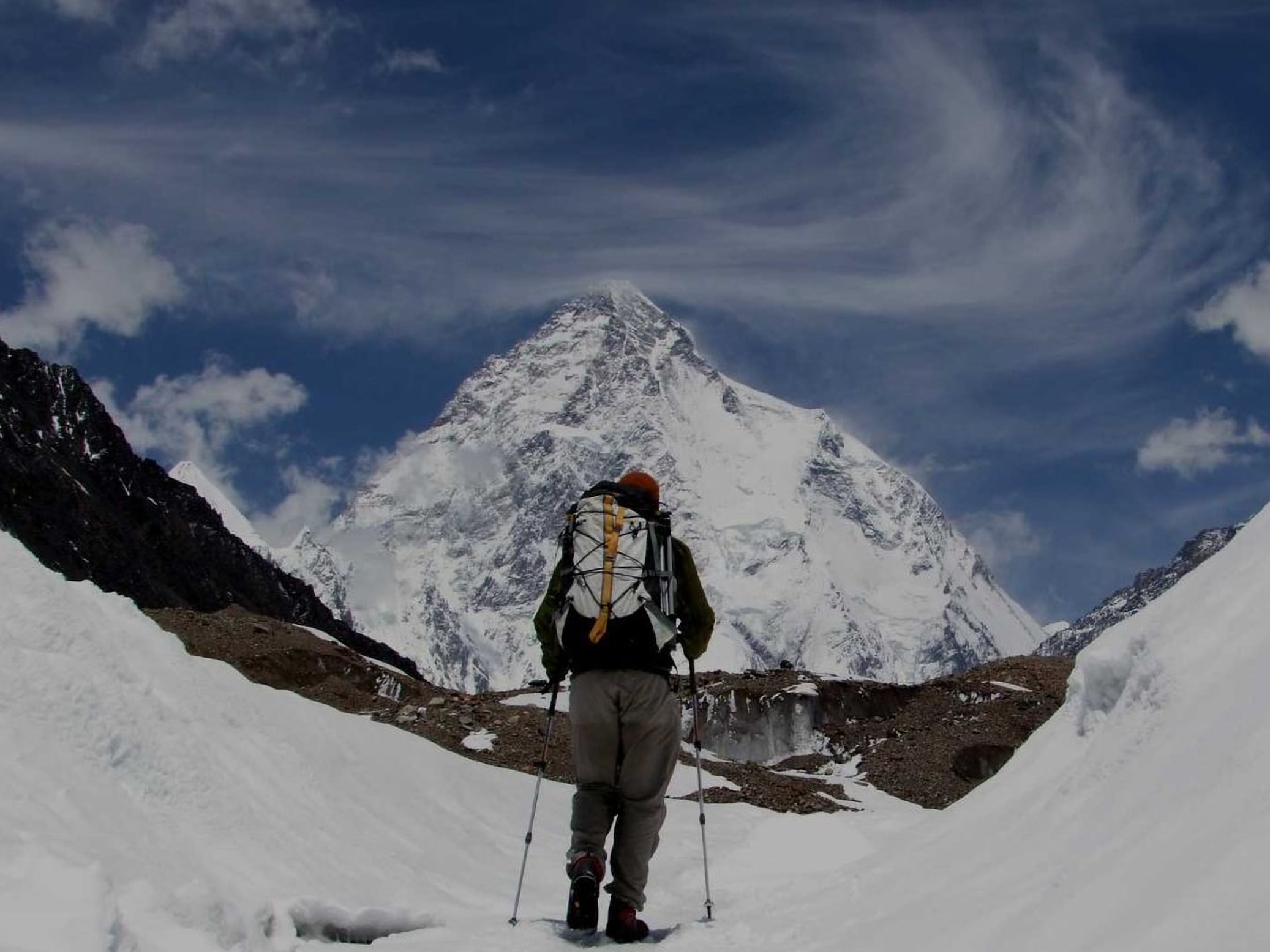 A trekker with a large backpack and walking poles crossing snow towards K2