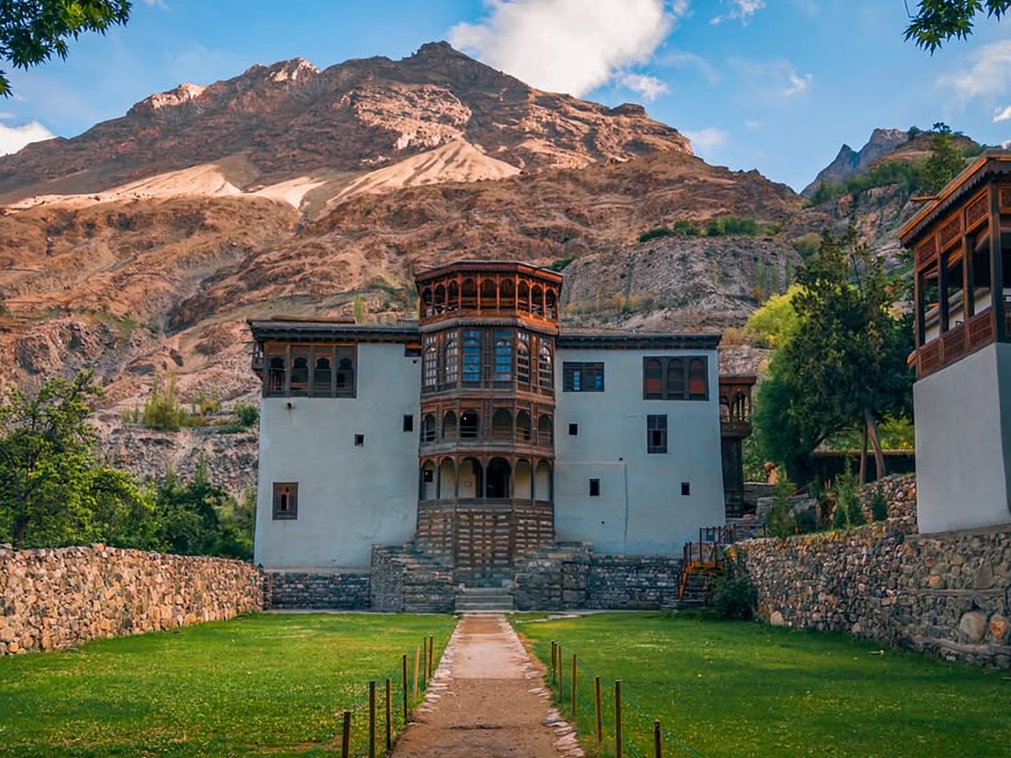 Khaplu Palace, a white palace with a carved wooden balcony tower, behind lawns and stone walls below rocky mountains