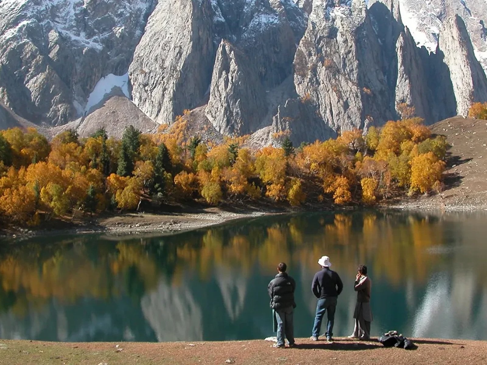Three walkers on the shore of Kutwal Lake, below golden autumn trees and granite spires reflected in the water