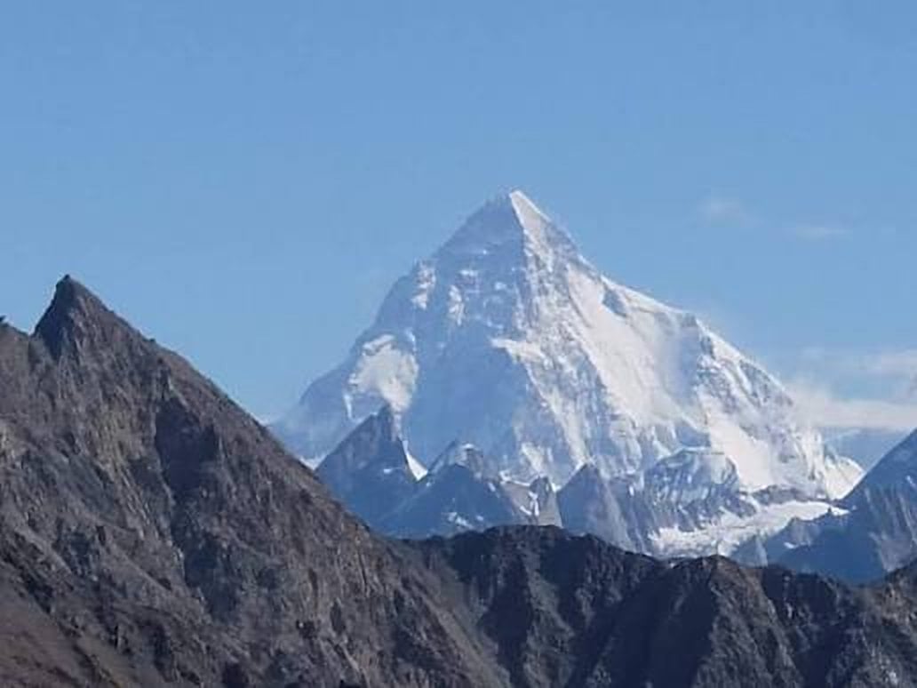 The snowy pyramid of K2 rising above dark rocky ridges under a clear blue sky, seen from Machulu La