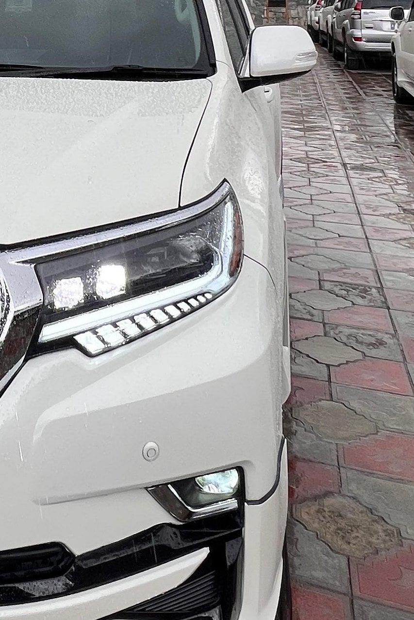 The headlight of a white Toyota Prado shining in the rain, with more Prados parked on a wet tiled drive