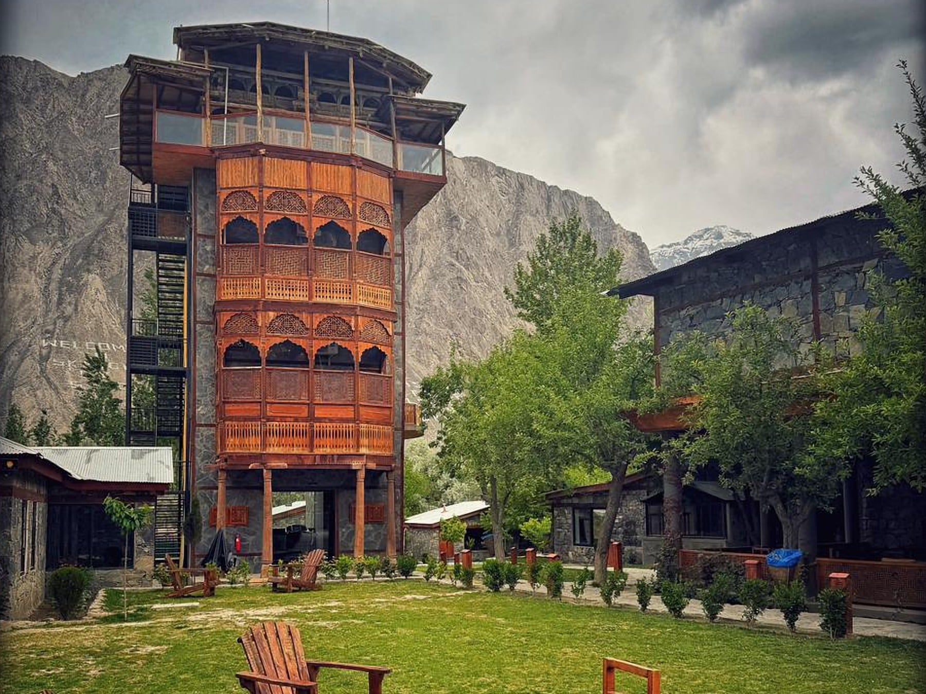 The carved wooden tower and lawns of PC Skardu, with wooden garden chairs and mountains under a cloudy sky