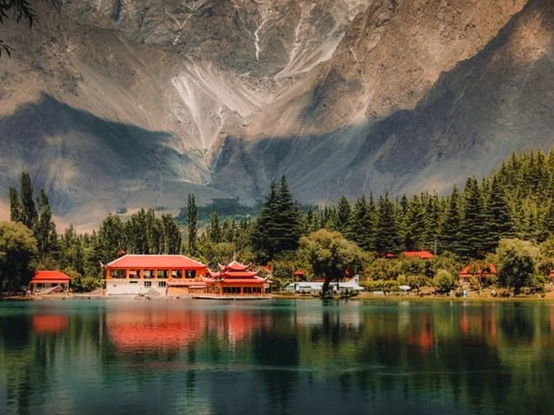 The red-roofed buildings of Shangrila resort reflected in Lower Kachura Lake, below pine trees and rocky mountains