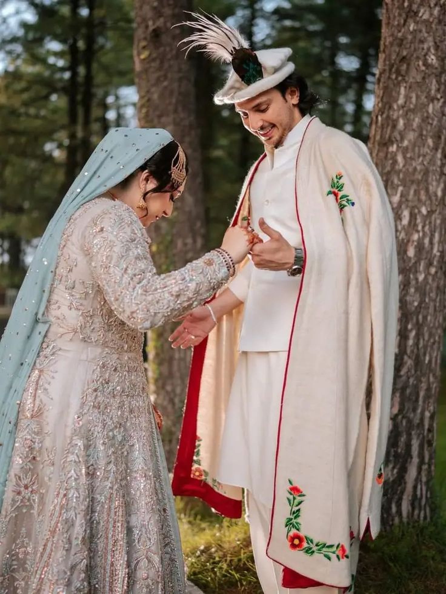A smiling bride and groom in traditional dress among pine trees: the bride in an embroidered silver gown and pale blue dupatta holds the groom's hand; he wears a cream embroidered robe and a feathered cap